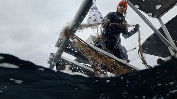A person in a wetsuit and red beanie stands on a metal platform above water, pulling up a tangled net or debris from the sea.
